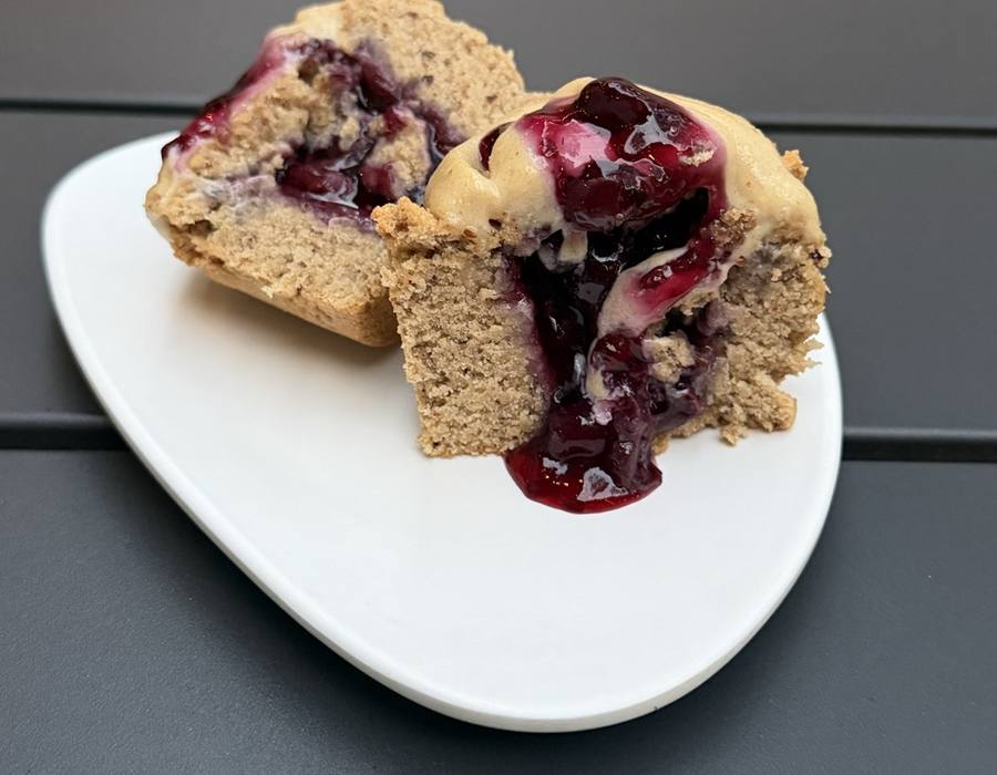A cupcake split open on a white plate, dark berry filling spilling from the centre and running down onto the plate, under a thick swirl of pale cashew creme